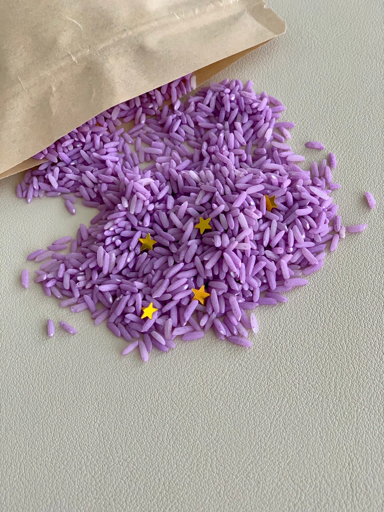 Small pile of dyed purple rice with gold star confetti spilling from brown paper bag on light surface