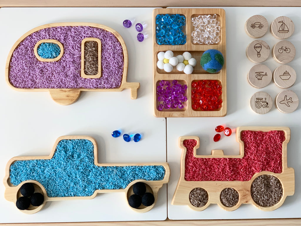 Wooden camper, truck, and train sensory trays filled with colored rice and gems on white surface with wooden vehicle stamps