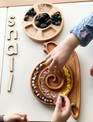 Cherry wood snail-shaped sensory tray with spiral grooves filled with colorful beads and hands arranging pieces on white table