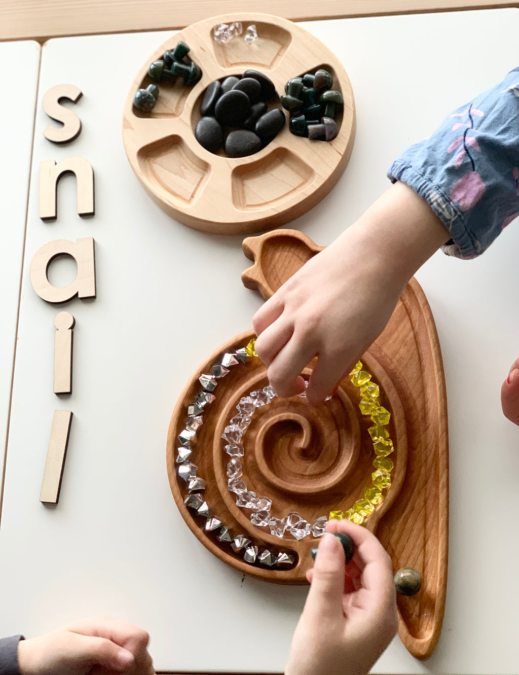 Cherry wood snail-shaped sensory tray with spiral grooves filled with colorful beads and hands arranging pieces on white table