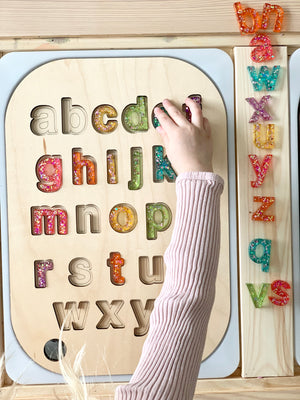 Child placing glitter resin lowercase letters into wooden alphabet puzzle with a drawstring bag and wooden board on table