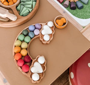 Wooden rainbow-shaped sensory tray with colorful felt balls and white felt hearts on tan surface