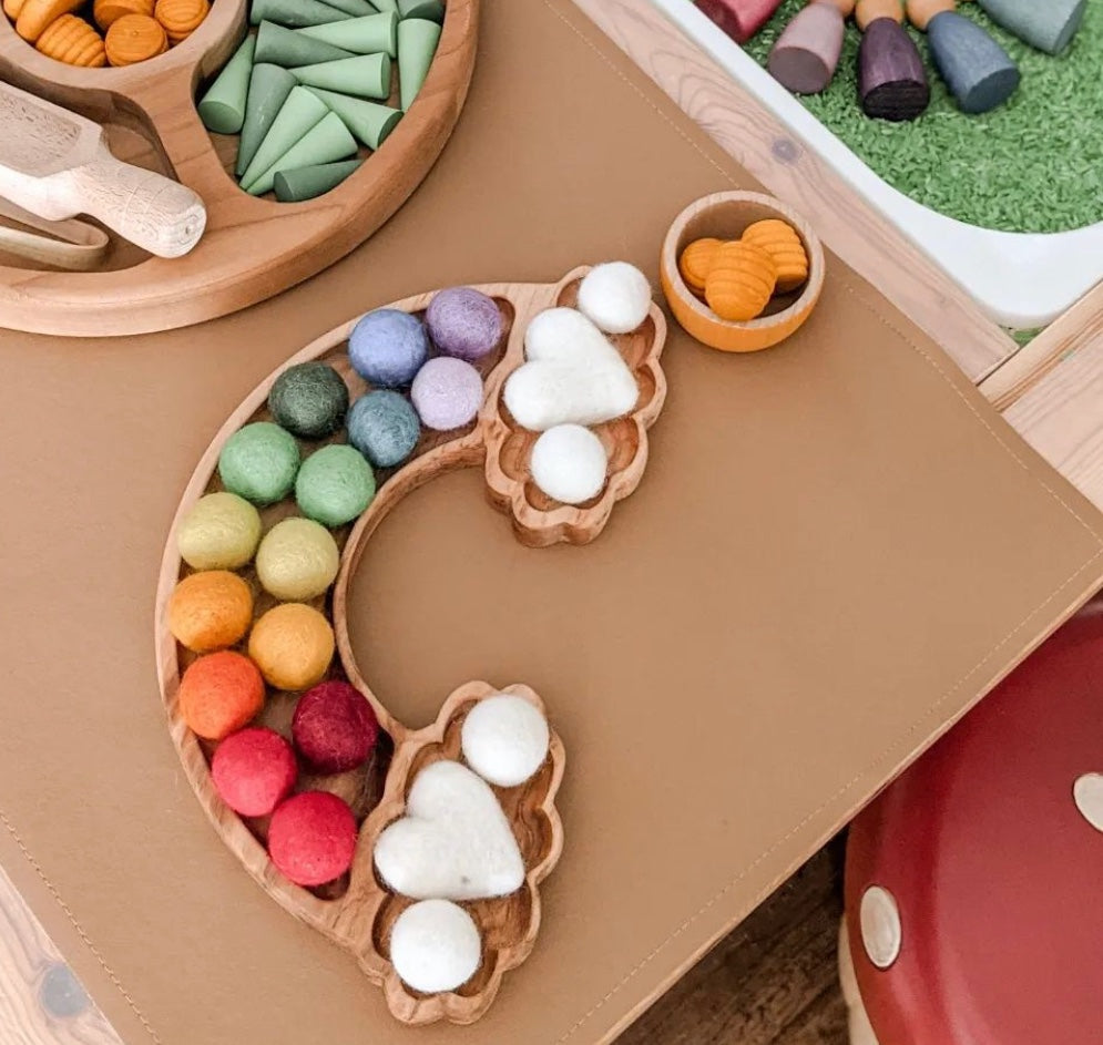 Wooden rainbow-shaped sensory tray with colorful felt balls and white felt hearts on tan surface