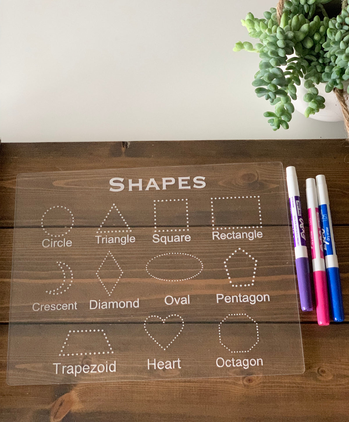 Clear acrylic tracing board with engraved shapes and names, and colorful dry erase markers on wooden surface