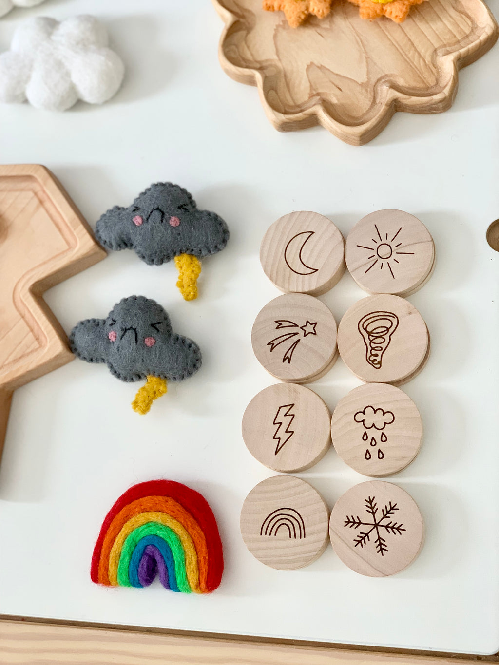 Natural wooden 2-inch discs engraved with weather symbols arranged on white surface with felt cloud and rainbow toys