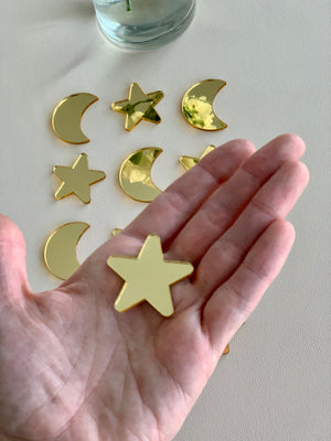 Hand holding shiny gold acrylic star shape with gold moon and star shapes scattered on white surface below
