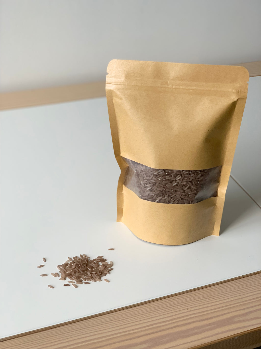Brown sensory filler grains inside a resealable kraft paper pouch with clear window on white surface with scattered grains