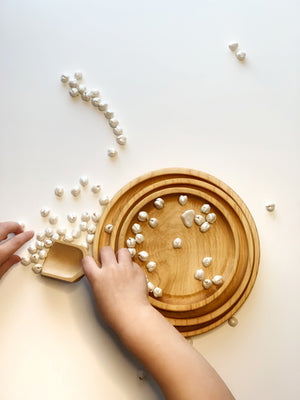 Small and medium nesting round wooden sensory trays with child’s hands playing with white beads on white surface