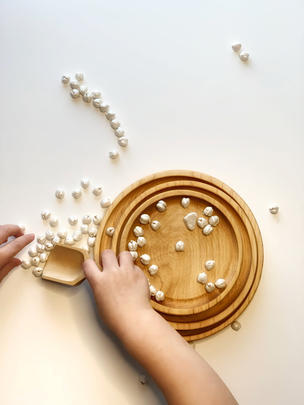 Small and medium nesting round wooden sensory trays with child’s hands playing with white beads on white surface