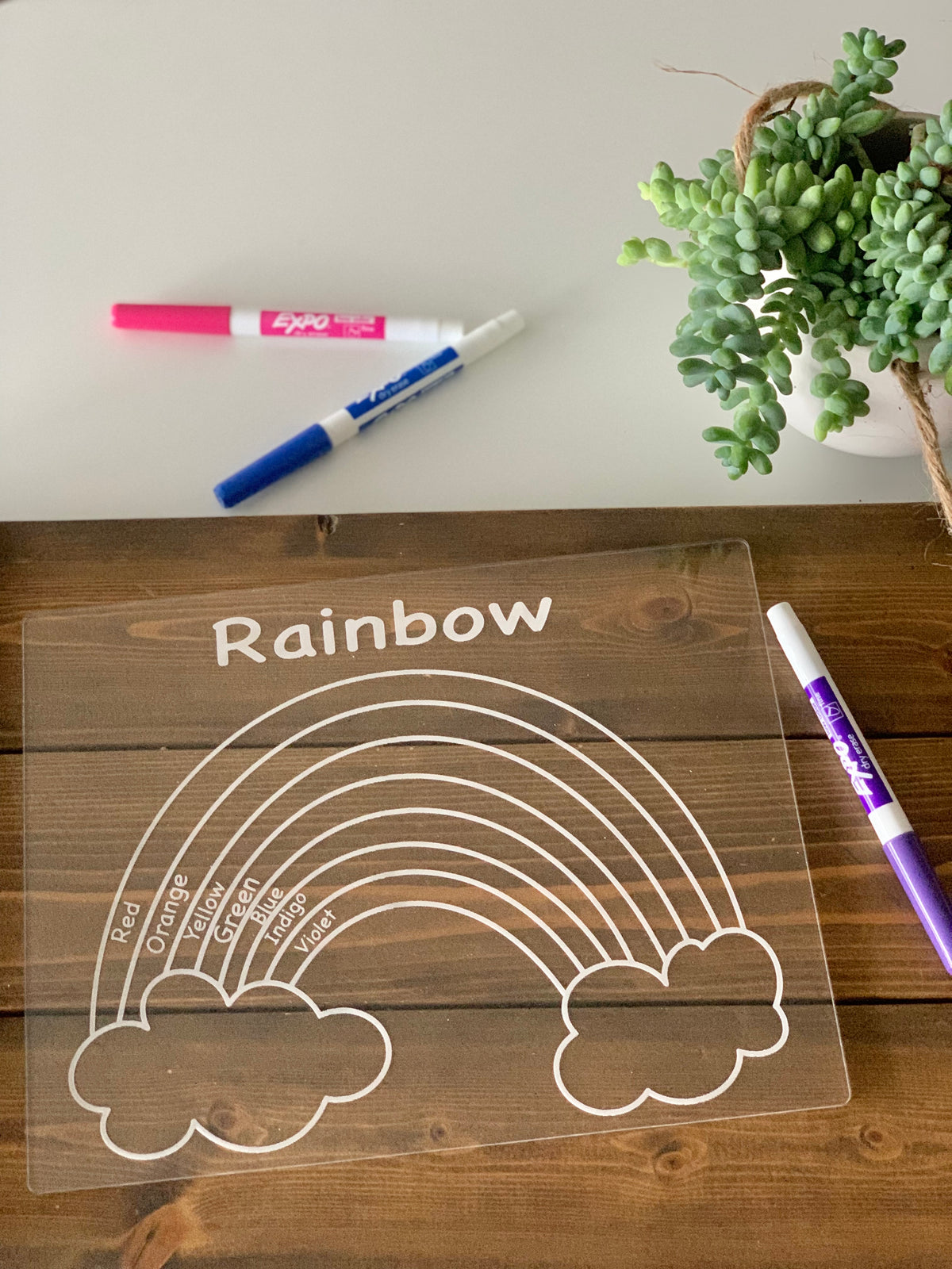 Clear acrylic dry erase board engraved with rainbow outline and color names on wooden table with markers and green plant