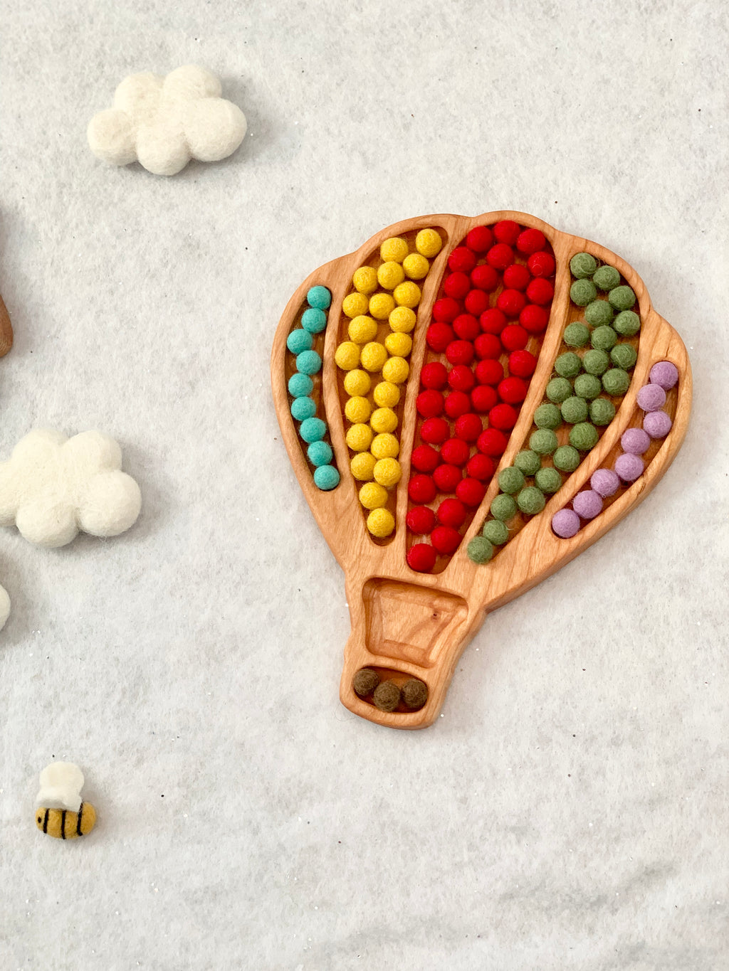 Wooden hot air balloon sensory tray filled with colorful felt balls surrounded by felt clouds and small felt bee on white surface