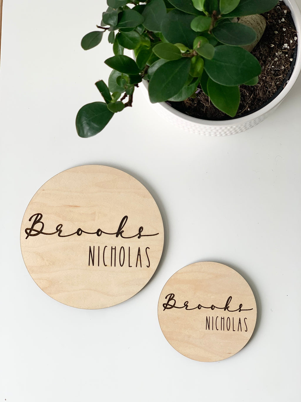 Two personalized round wooden baby name announcement discs with "Brooks Nicholas" engraved near green potted plant on white surface