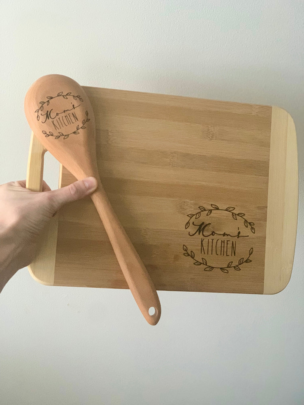 Hand holding personalized wooden spoon engraved with "Mama's Kitchen" next to matching engraved bamboo cutting board