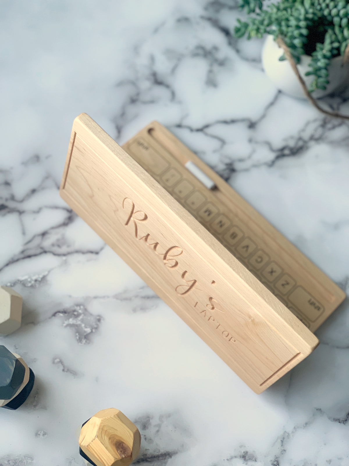 Wooden toy laptop with engraved name and chalkboard screen on marble surface with decorative plants and toys