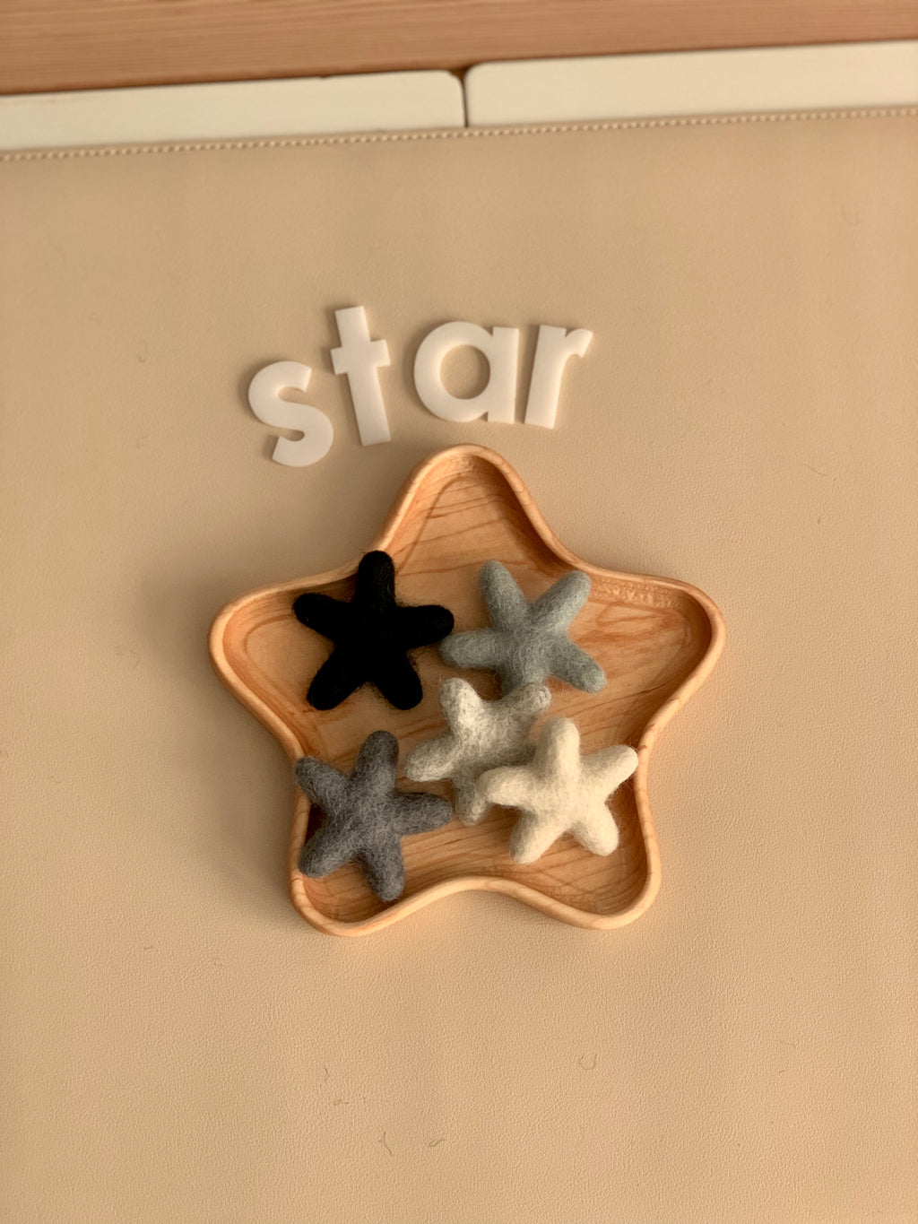 Small handmade neutral felt stars in black, gray, and white arranged inside wooden star-shaped tray on beige surface