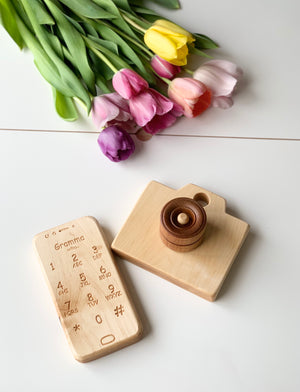 Natural wooden toy camera with engraved rotating lens and matching wooden toy phone with engraved buttons on white surface with tulips