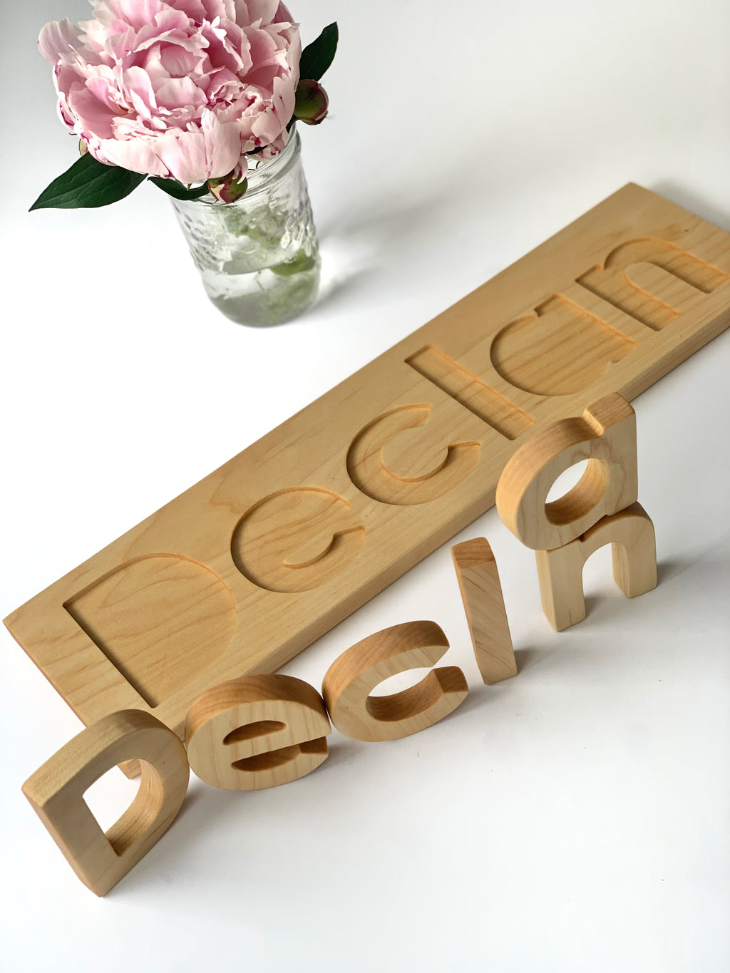 Personalized hardwood name puzzle with natural wood finish next to pink peony in glass jar on white surface