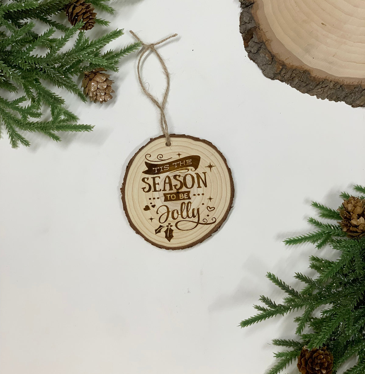 Round wood burned Christmas ornament with "Tis the Season to be Jolly" text and twine hanger on white background with pine branches