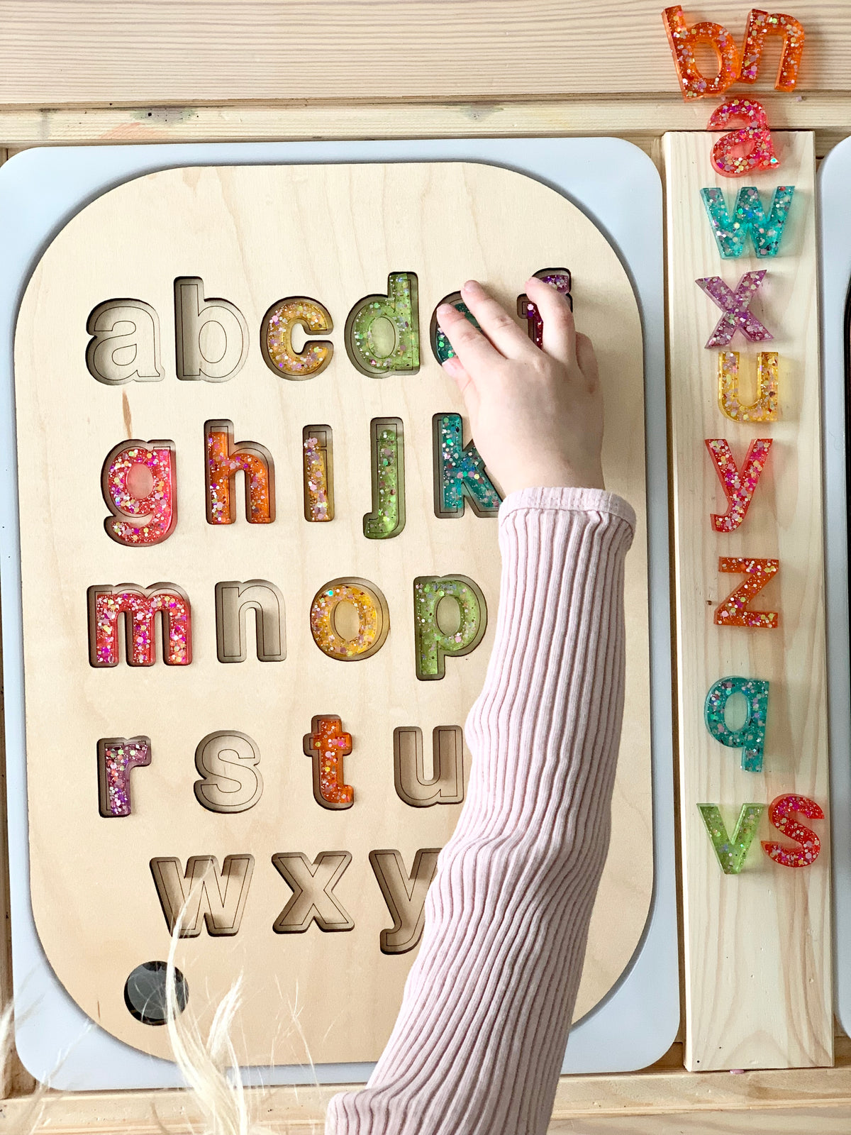 Wooden lowercase alphabet puzzle insert with colorful resin letters and child's hand placing letter on Flisat table top