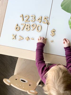 Maple wooden numbers and math symbols arranged on white table with child’s hands and bear-themed chair visible