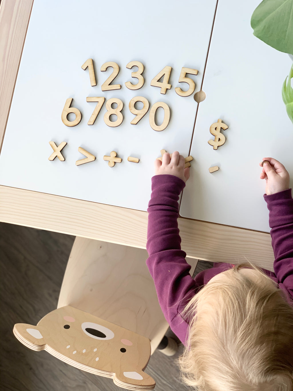 Maple wooden numbers and math symbols arranged on white table with child’s hands and bear-themed chair visible