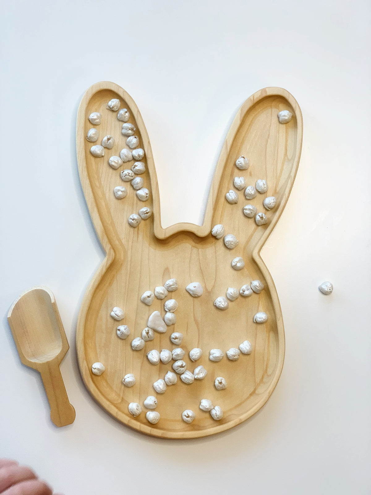 Large wooden bunny-shaped sensory tray with scattered small white textured beads and matching wooden scoop on white surface