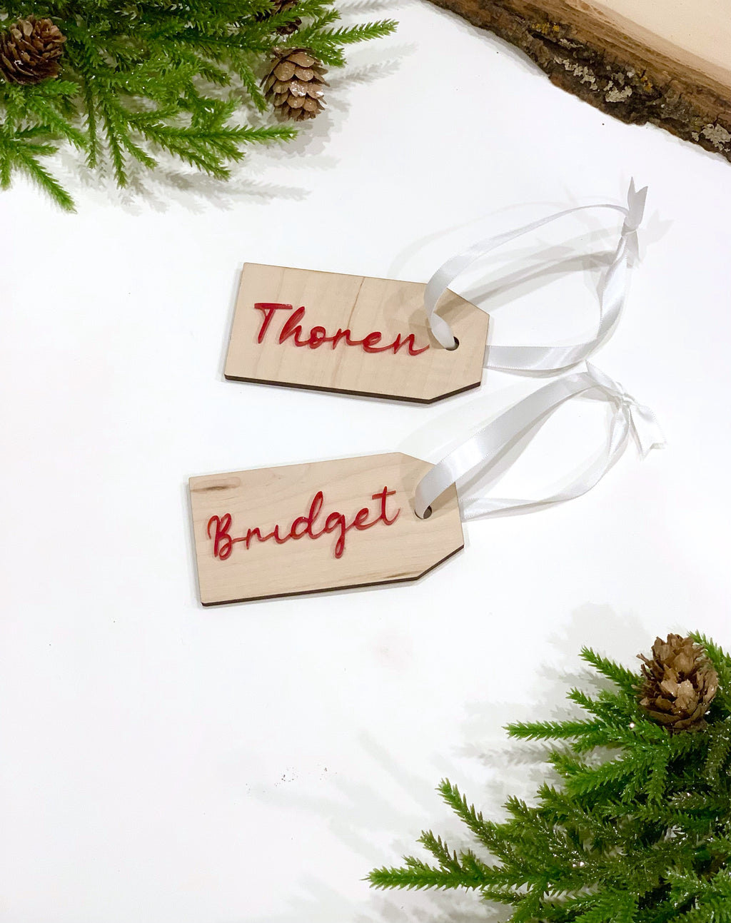 Two custom wooden Christmas gift tags with red cursive names and white ribbon on white background with pine branches and cones