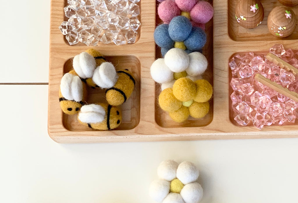 Natural wooden rectangle tinker tray with multiple compartments holding felt balls and clear acrylic gems on white surface