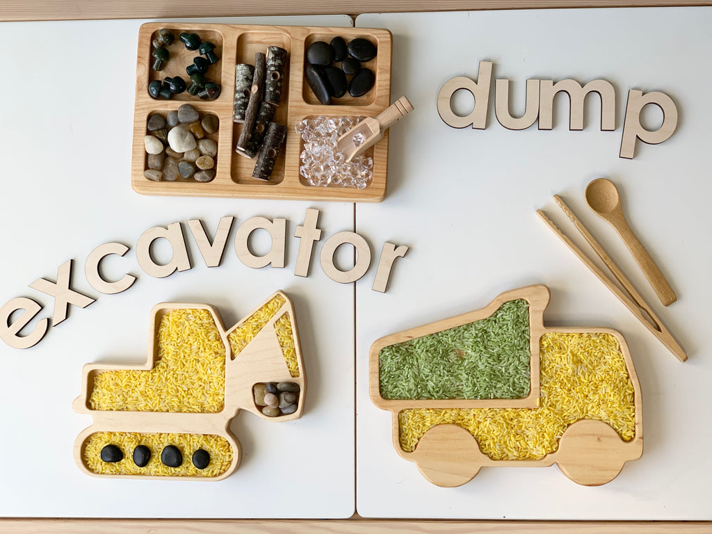 Wooden excavator and dump truck sensory trays filled with colored rice and small rocks with wooden letters and utensils on white surface