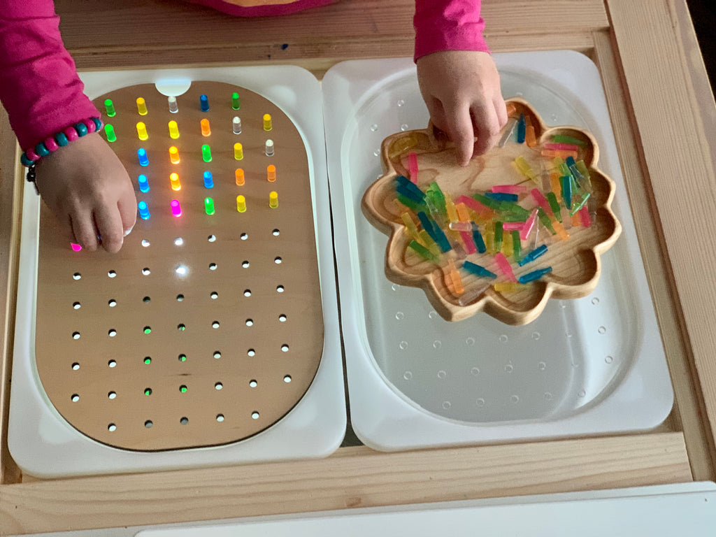 Child’s hands placing colorful translucent pegs into a small hole geometric light board on wooden table surface