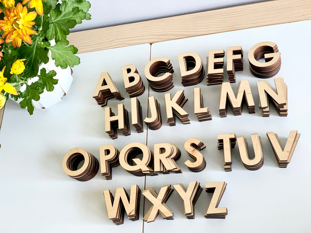 Uppercase wooden alphabet letters set made of maple wood arranged on white surface near yellow flowering plant