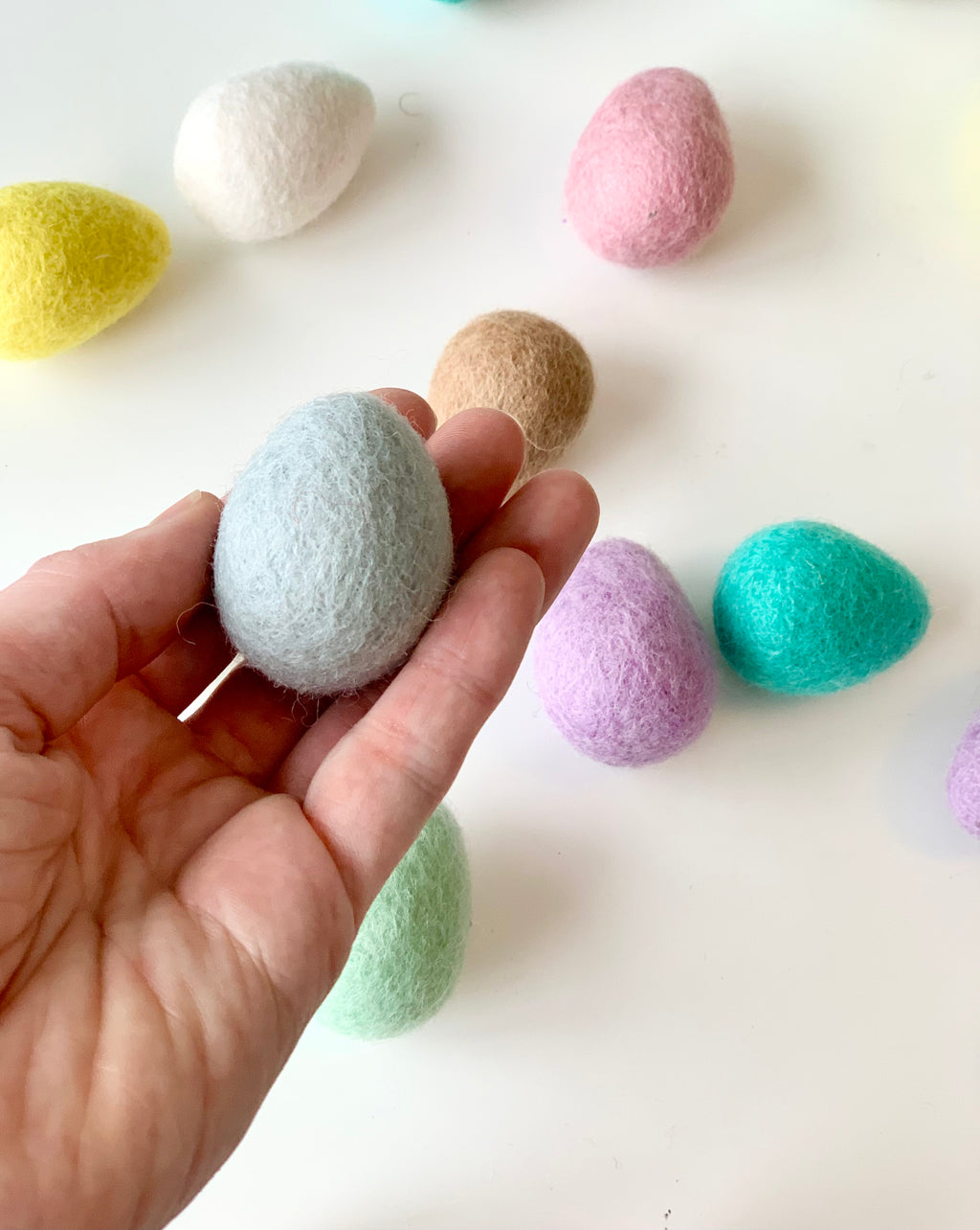 Hand holding one light blue felt Easter egg with other pastel-colored felt eggs on white surface