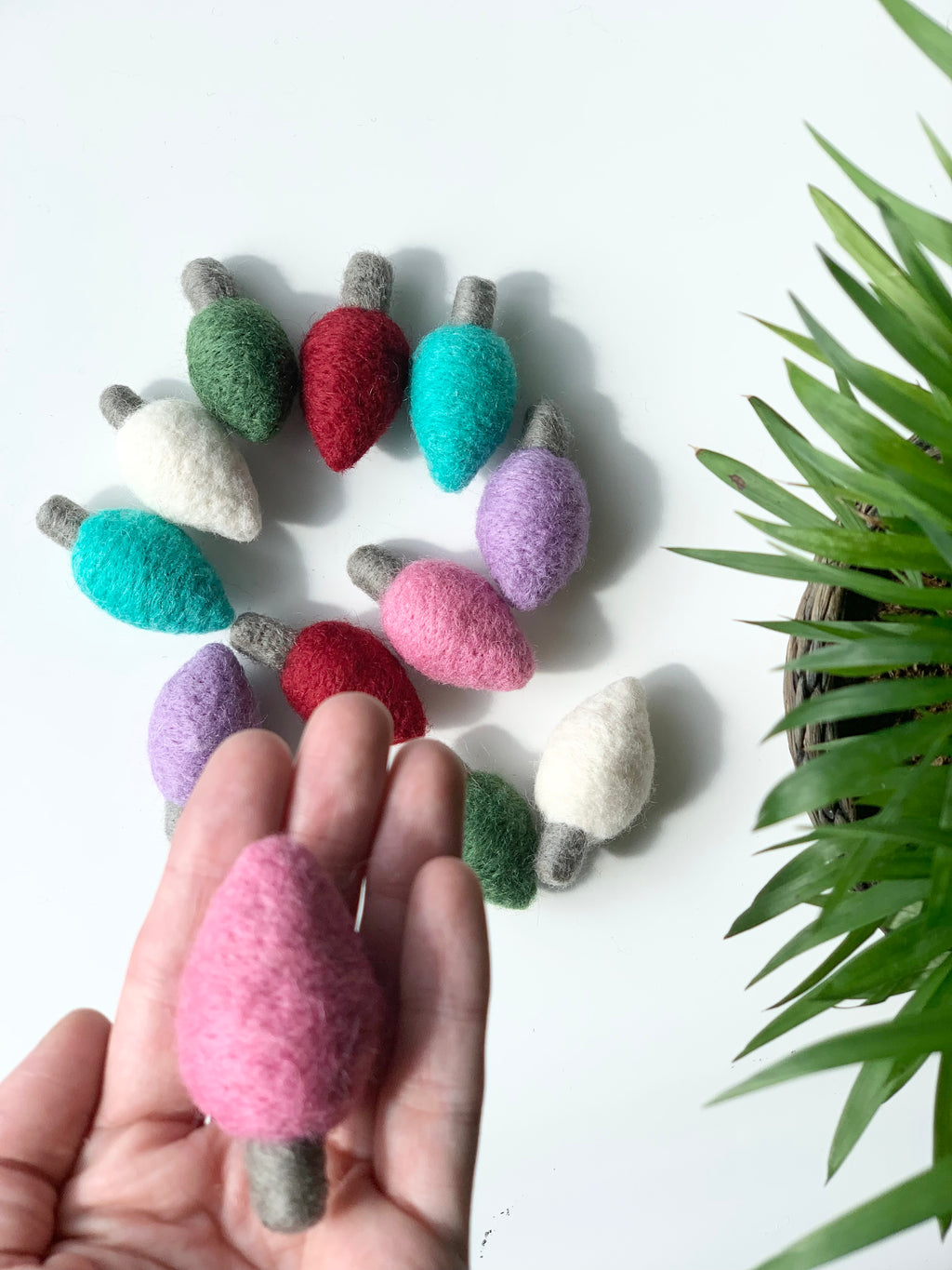 Hand holding one pink felt light bulb with more colorful felt bulbs arranged in a circle on white surface near green plant
