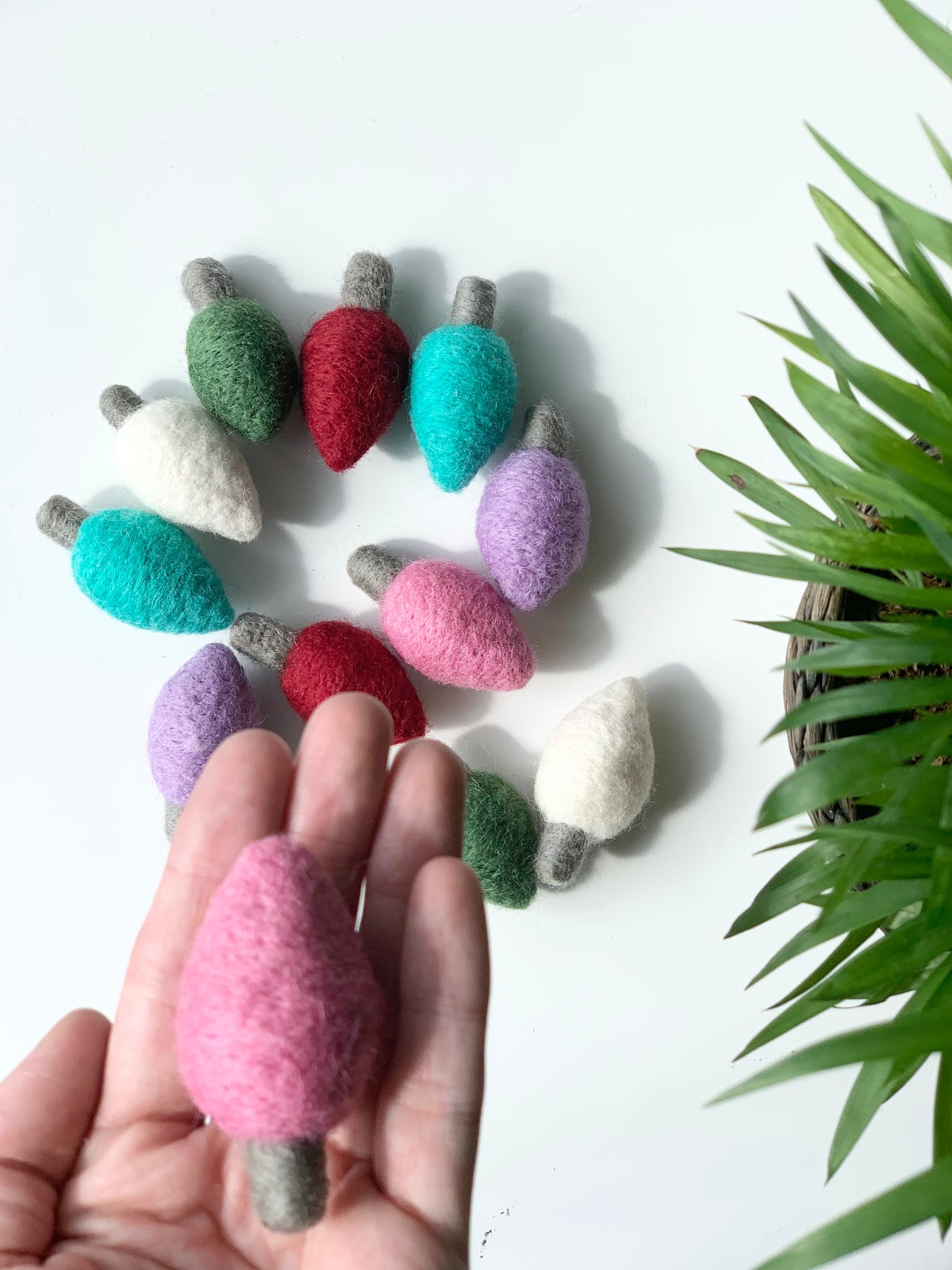 Hand holding one pink felt light bulb with more colorful felt bulbs arranged in a circle on white surface near green plant