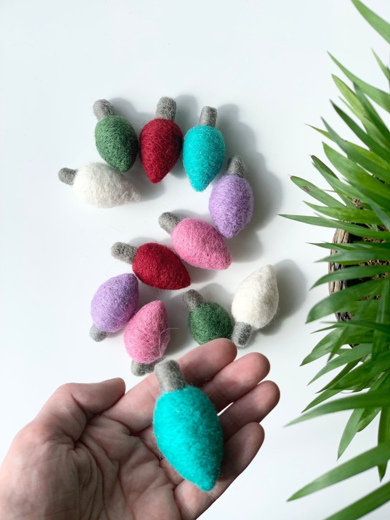 Assorted felt Christmas lightbulbs in various colors including robin's egg blue held in hand on white background with green plant