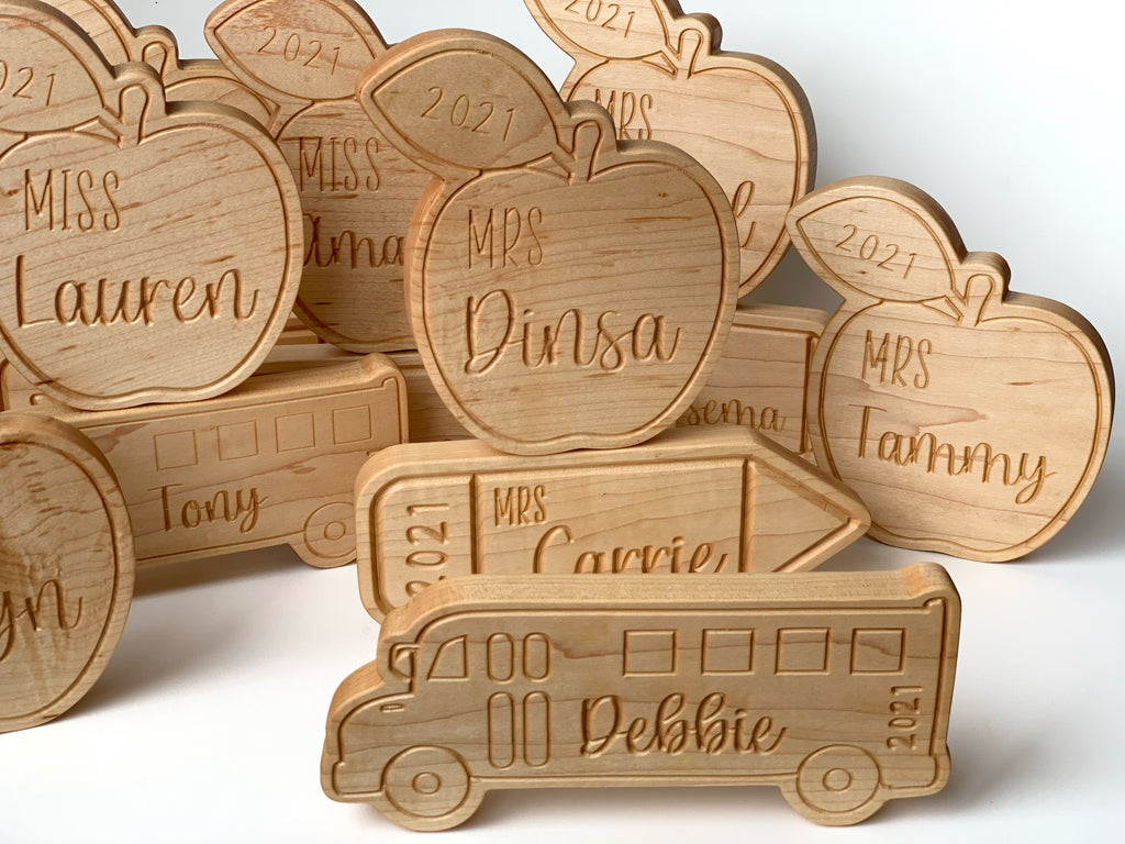 Personalized natural wood teacher name plaques shaped like apples, pencils, and buses engraved with names and year 2021