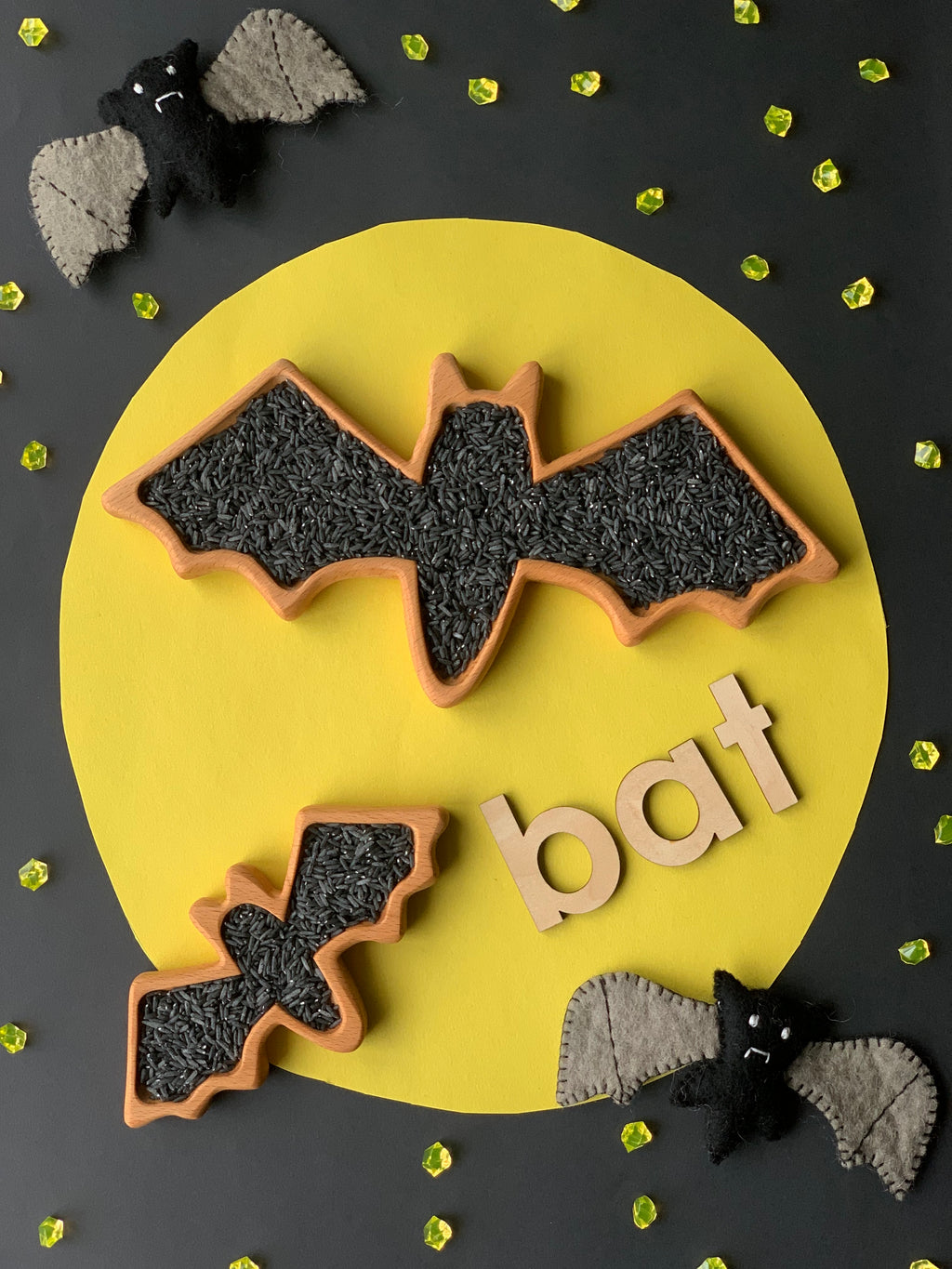 Bat-shaped wooden sensory trays filled with black rice on yellow circle background with felt bats and wooden letters spelling bat