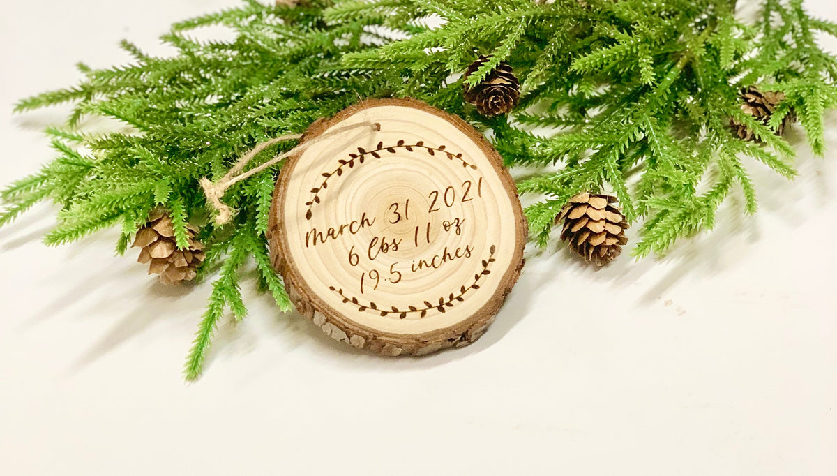 Wood burned wooden baby’s first Christmas ornament with birth details surrounded by pine branches and pinecones