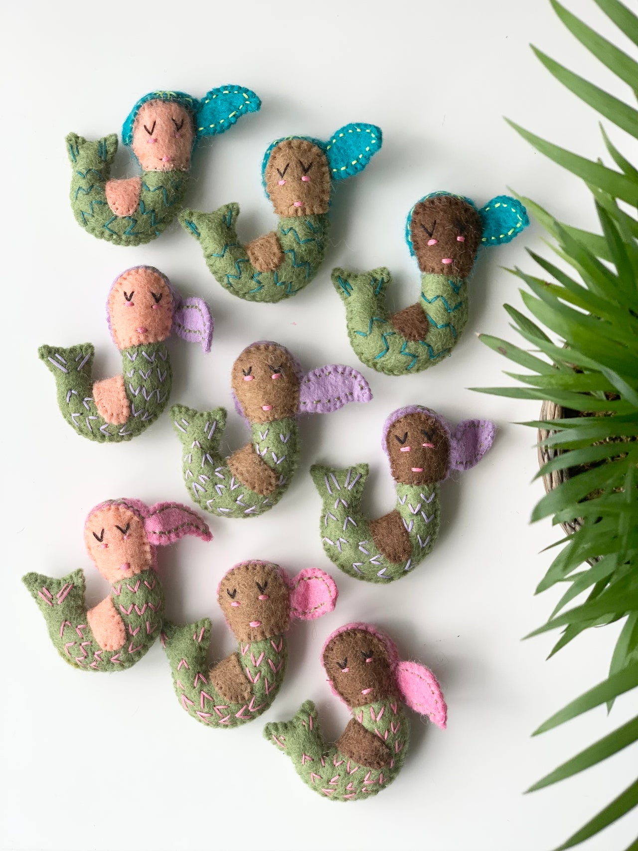 Collection of small handmade felt mermaids in turquoise and pink fins with stitched details on white background near green plant leaves