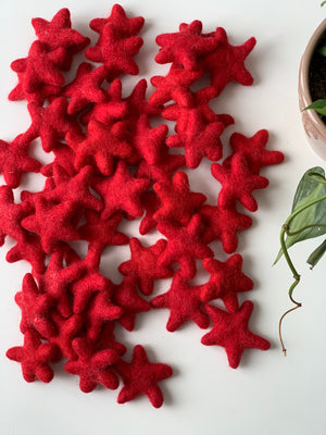 Multiple handmade red felt stars made from pure wool scattered on white surface near green potted plant