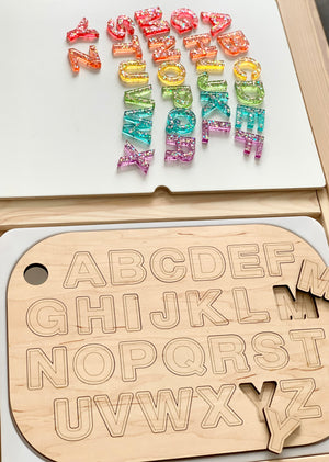 Wooden uppercase alphabet puzzle board with colorful resin letters laid out above on white surface and wood table edge