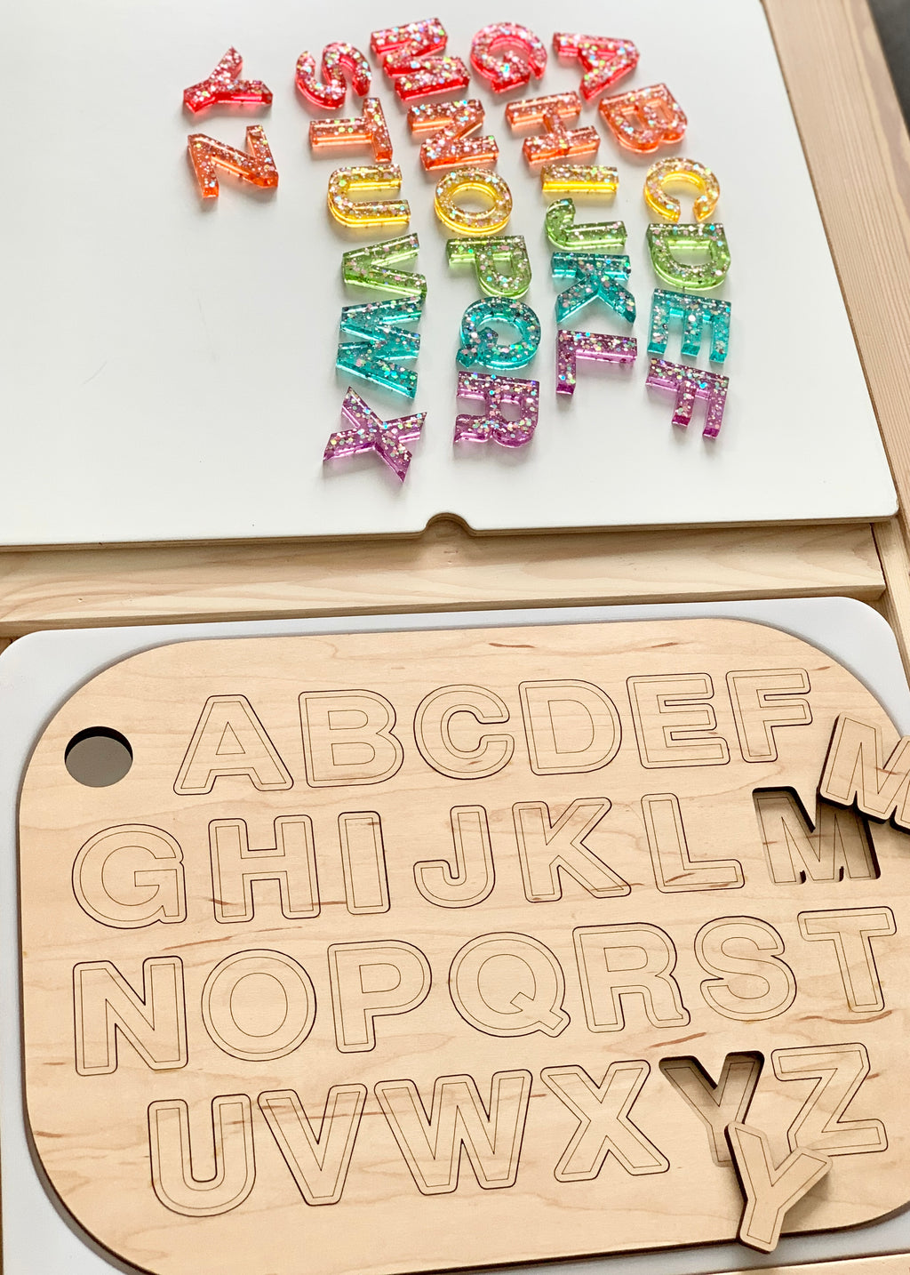 Wooden uppercase alphabet puzzle board with colorful resin letters laid out above on white surface and wood table edge