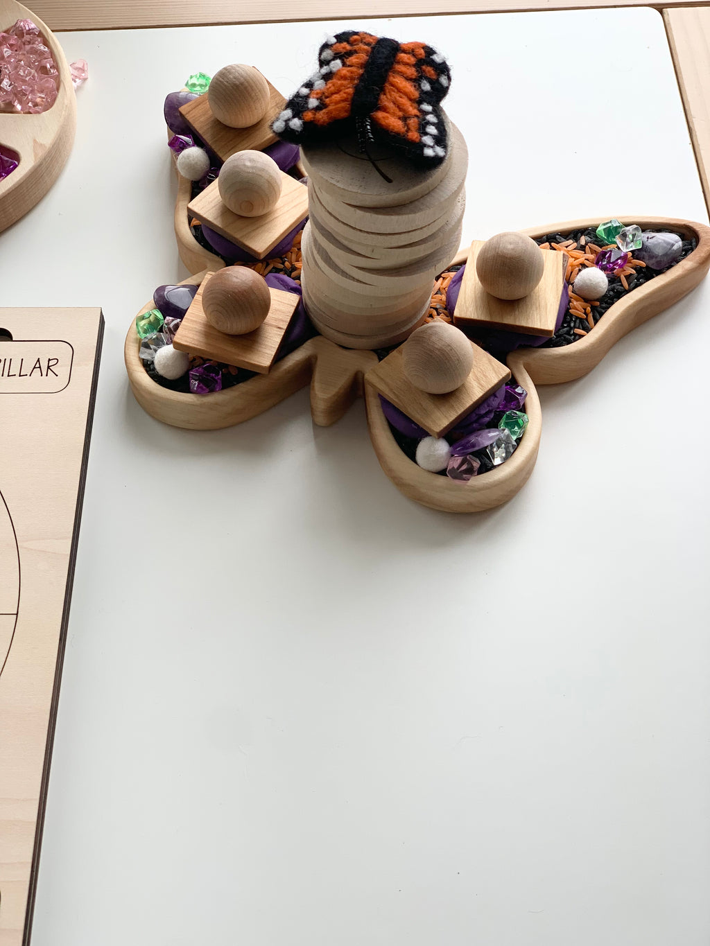 Butterfly-shaped wooden sensory tray with wooden blocks, beads, and colorful textured materials on white surface