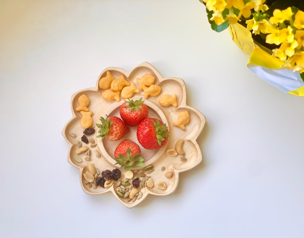 Sunflower-shaped wooden sensory tray with strawberries, goldfish crackers, nuts, and dried fruit on white surface with yellow flowers nearby