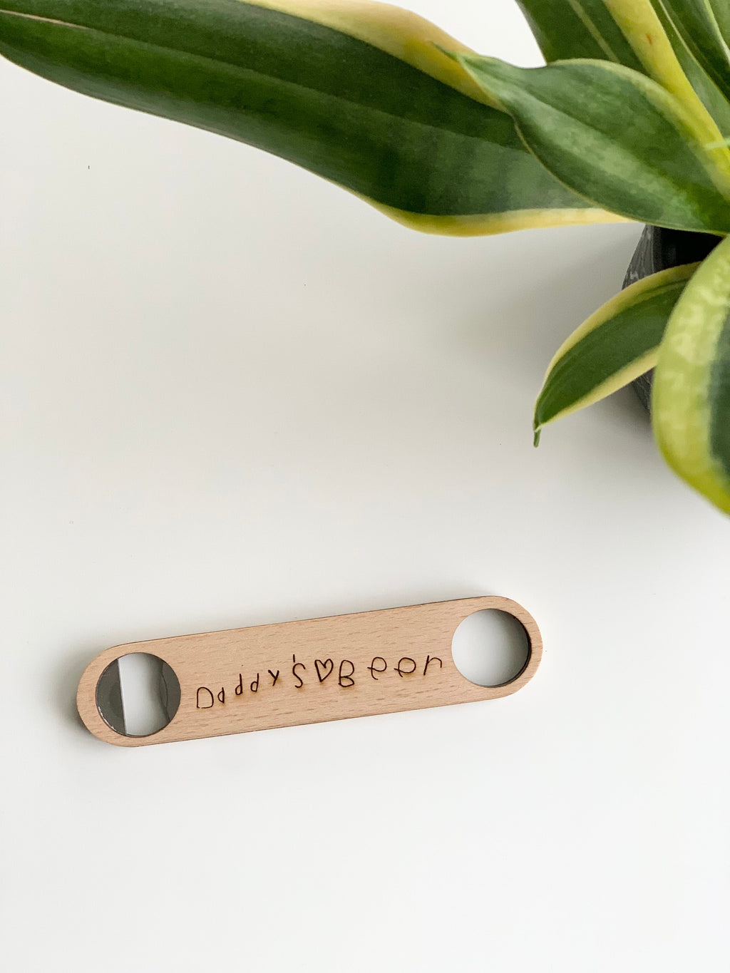 Custom wooden bottle opener with handwritten style engraving on light natural wood background near green plant leaves