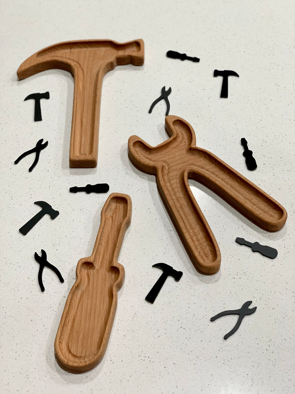 Wooden sensory tray shaped like hammer, pliers, and screwdriver with black tool-shaped cutouts on white surface