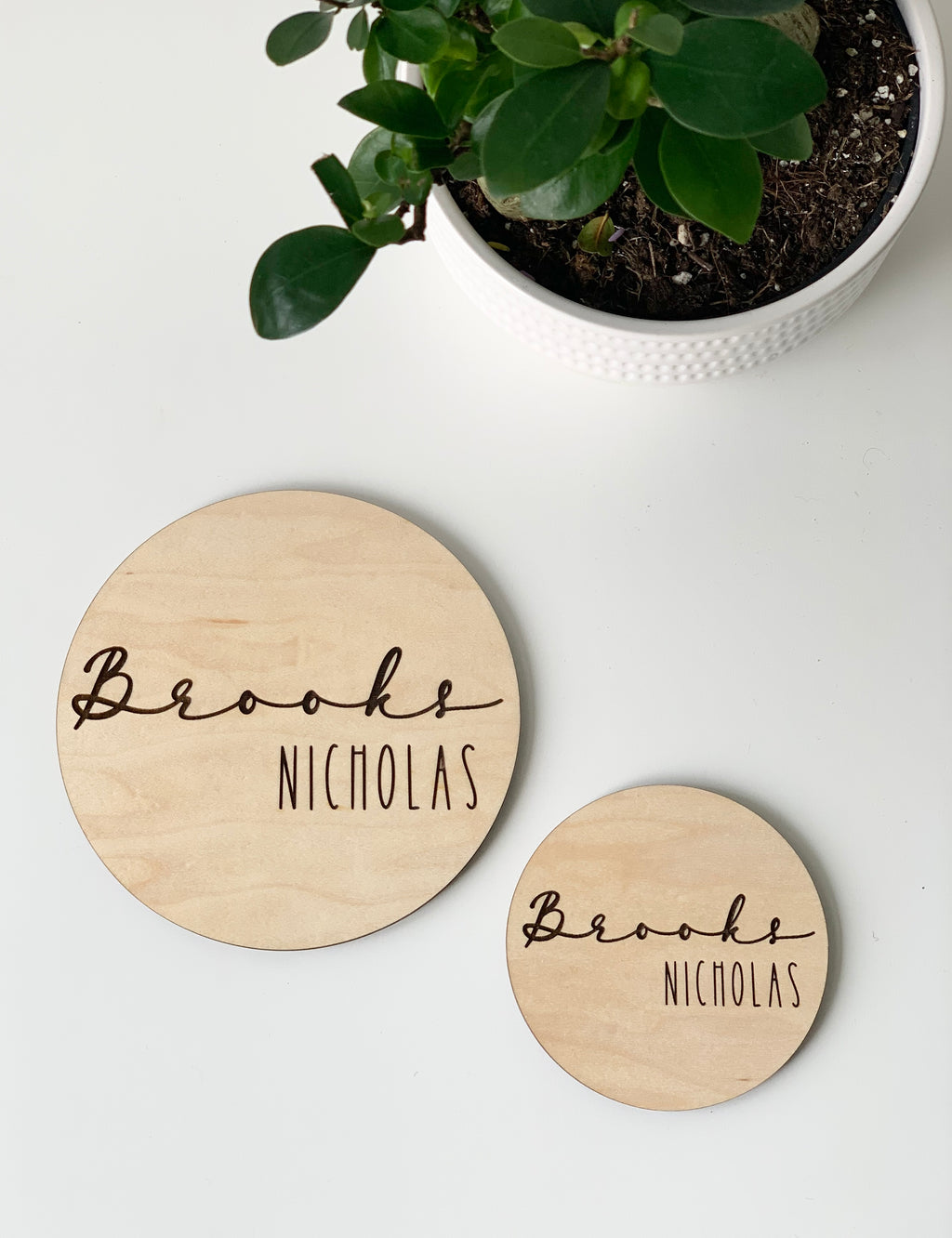 Two personalized round wooden name announcement discs engraved with the name Brooks Nicholas on white surface next to green plant