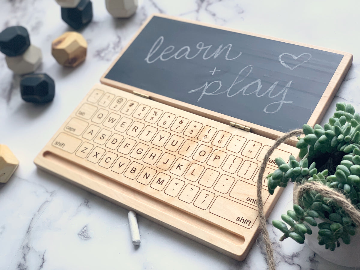 Wooden laptop toy with engraved keyboard and chalkboard screen displaying handwritten learn plus play on marble background