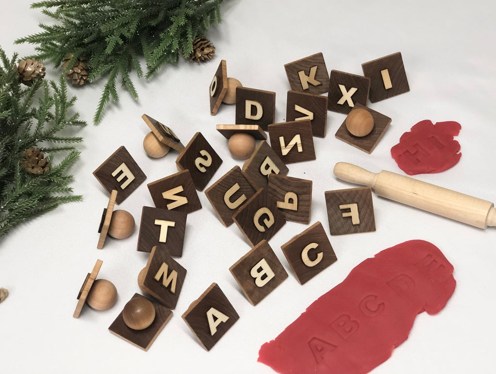 Wooden alphabet play dough stampers with round handles scattered near a rolling pin and red play dough on white surface
