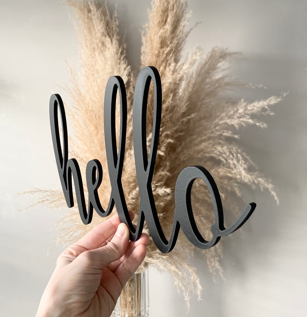 Hand holding custom black wooden word cutout saying hello with pampas grass background
